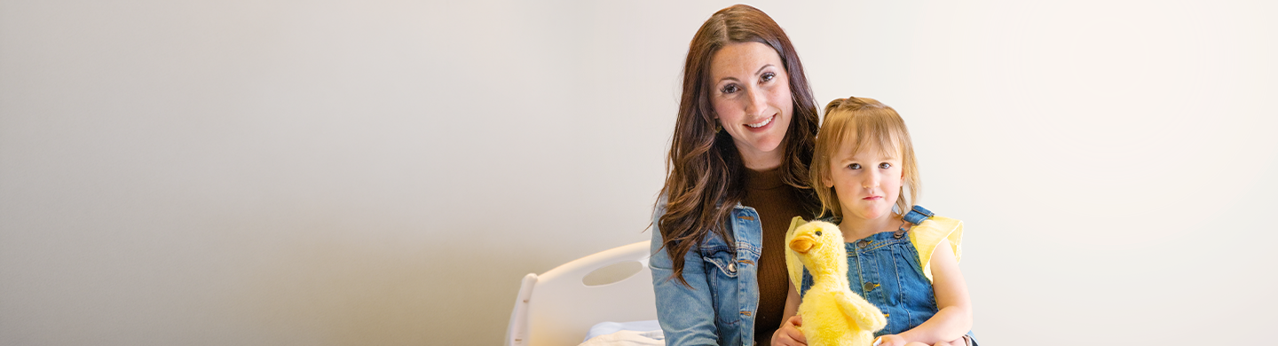 Adult sitting on a hospital bed with a child holding a yellow plush duck, both facing the camera against a plain background.